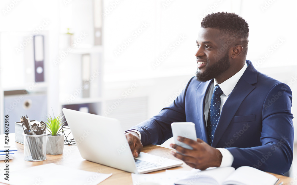 Dreaming young entrepreneur working with laptop in office, holding smartphone