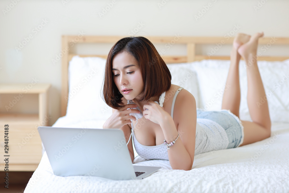 side view shot of beautiful adult asian woman in white dress lying on bed in bedroom while using computer and listening music from headphone side view shot of beautiful adult asian woman in white dress lying on bed in bedroom while using computer and listening music from headphone