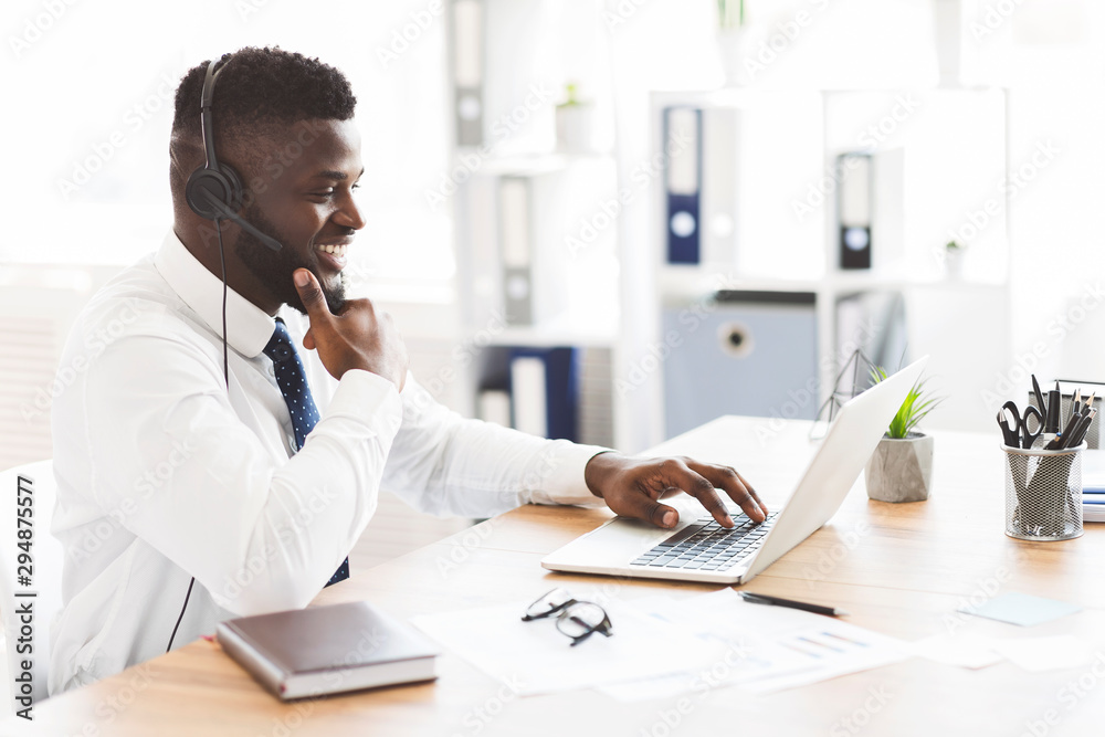 Cheerful young worker with headset sitting in front of laptop Cheerful young worker with headset sitting in front of laptop