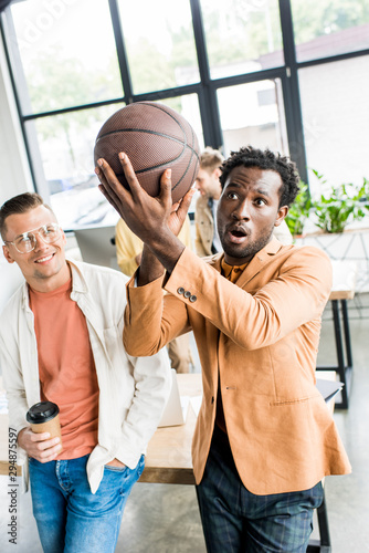 african american businessman playing with volleyball ball while standing near colleague holding coffee to go in office