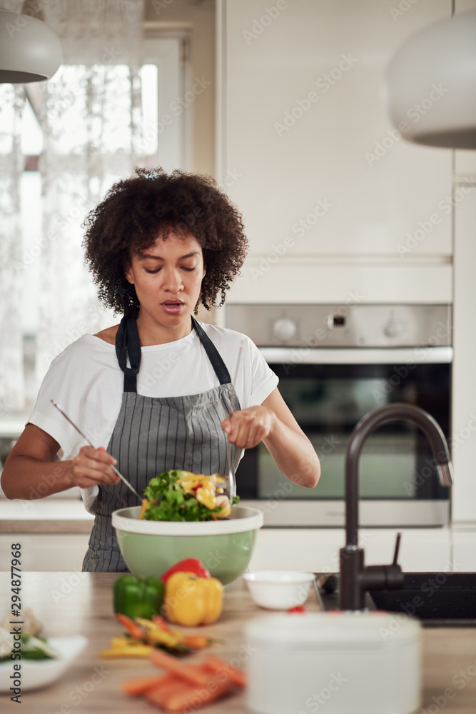 Attractive mixed race woman in apron and with curly hair mixing vegetables in bowl while standing in kitchen at home. Attractive mixed race woman in apron and with curly hair mixing vegetables in bowl while standing in kitchen at home.