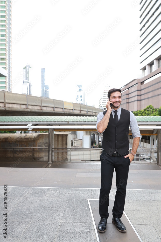 full length successful caucasian businessman in black suite who standing in downtown, he using cell phone with happy smiling face with city and skytrain background