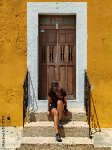 Woman sitting in a typical colonial facade in yellow with a wooden door, Izamal (Mexico)