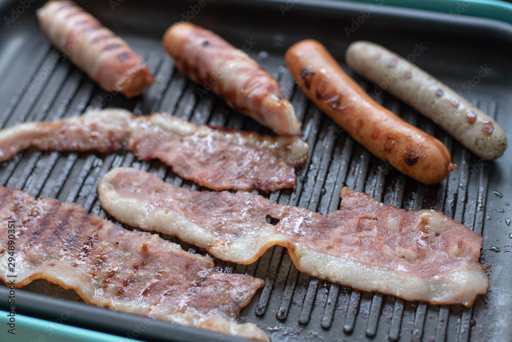Assorted sausages and bacon being cooked on an electrical griddle plate.
