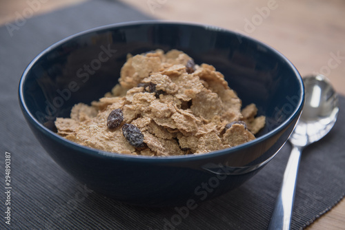 A dark blue colored bowl of delicious corn flake cereals with raisins, milk and a spoon beside a wooden surface.
