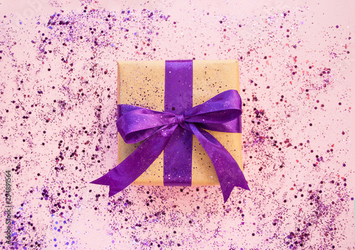 Giftbox tied with purple color ribbon on pastelpink background with glitter. Flat lay style.