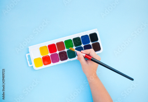 Child's hands holding bpain brsh and watercolor paints on blue background with copyspace. Flat lay style. Back to school concept. photo