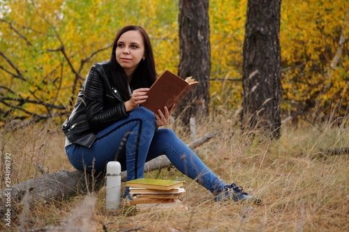 A young beautiful woman with long dark hair in casual clothes is sitting on a log and reading a book in the autumn forest.
