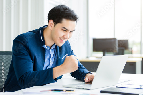 Happy of asian young businessman see a successful business plan on the laptop computer and pen on wooden table background in office,business expressed confidence embolden