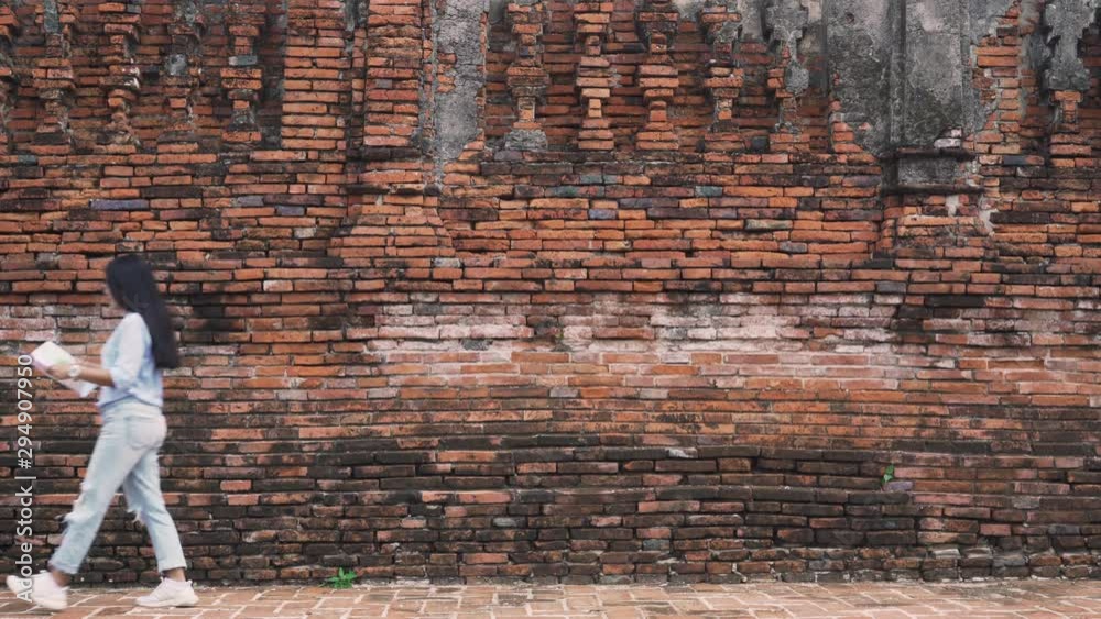 Side view of a young Asian female tourist walking past an old red brick wall in an ancient city while looking at a map - Millennial girl on holiday exploring attractions in the city - tourist concept