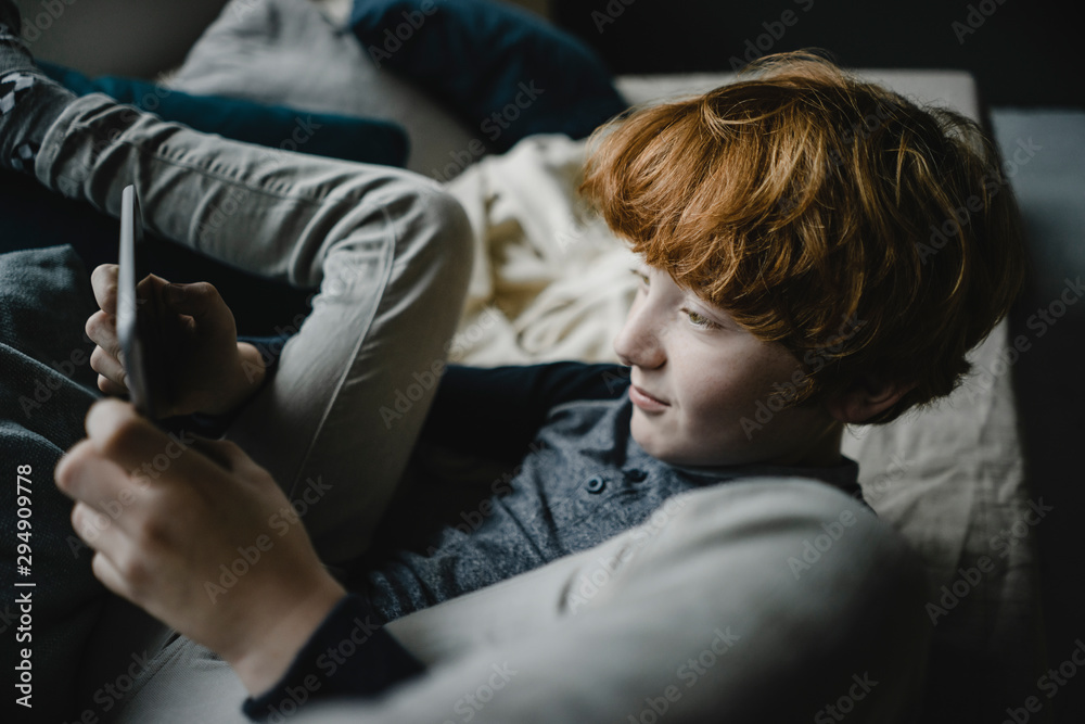 Redheaded boy lying on couch using digital tablet Redheaded boy lying on couch using digital tablet
