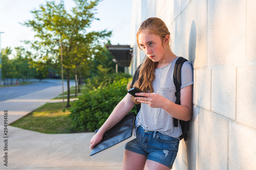 Depressed/Sad teen girl leaning against high school wall during sunset while wearing a backpack and holding binders/smartphone.