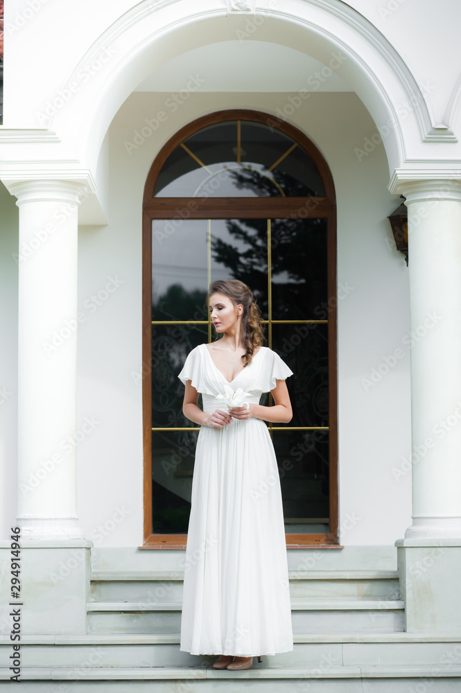 Fototapeta premium Young elegant woman with wedding bouquet in a light long white dress before ceremony