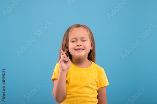 Close up emotional portrait of a funny girl with with eyes closed suggesting a desire and showing hand with crossed fingers against blue background with copy space in studio