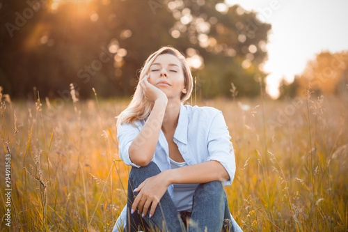 Young woman sitting on dry grass under sunlight. Eyes closed