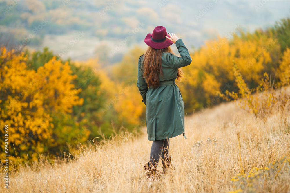 Beautiful young stylish girl in a coat walks in the autumn in the park. The girl is dressed in a green coat and a red hat. Beautiful evening. Autumn fashion. Lifestyle. High fashion portrait. 