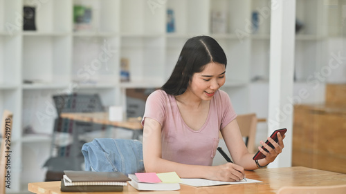 Young woman reading on smartphone and writing with note paper in libarary room. photo