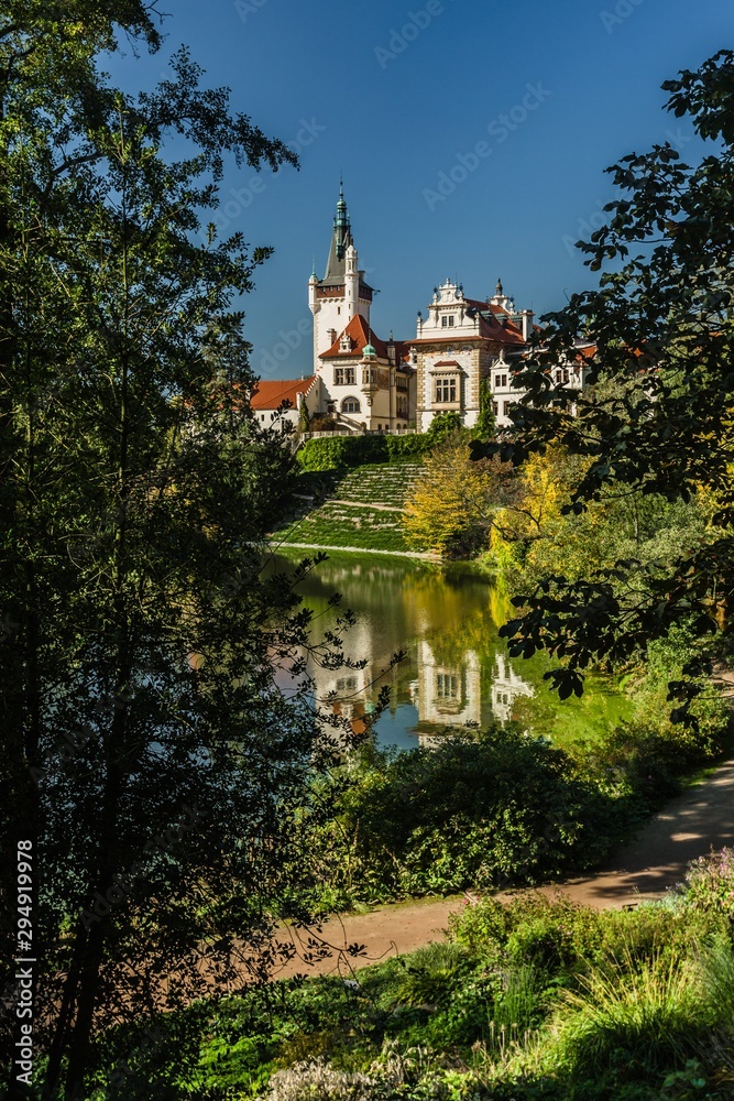 Fototapeta premium Pruhonice, Czech Republic - October 7 2019: Scenic view of famous romantic castle over a lake with its reflection in water, standing on hill surrounded with green trees. Sunny autumn day with blue sky