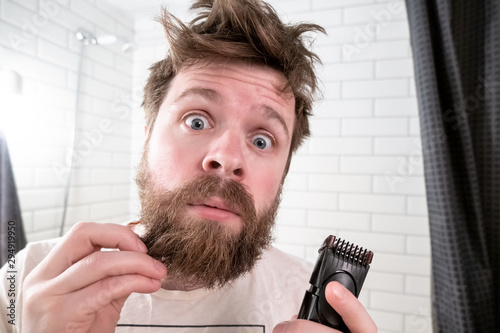 Shocked man holds a trimmer in hands and looks with wide eyes at his reflection in the mirror. He is terrified of his long beard and shaggy hairstyle.