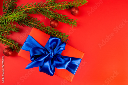 Red gift box with a blue bow near fir branches on a red background. a Christmas gift