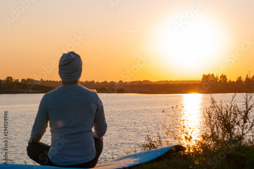 A young man sits on a lake at sunset, doing yoga. Sits in the pose of ardhapadmasana, padmasana. Balance, harmony, balance, concentration, relaxation.