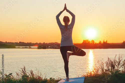 A young girl is standing on the lake at sunset, doing yoga. Stands in a pose of tree a Sathi Yoga. Balance, harmony, balance, concentration, relaxation. photo