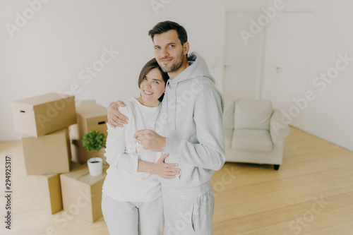 Millennial couple with plesant smiles, hug and hold key from own apartment, buy their first home, moved in shared house, stand in empty room with carton boxes in background. Ownership concept photo