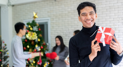 close up young handsome latin man smiling with holding red gift box in living room with group of asian friends decorating pine tree and talking to prepare christmas party celebration tonight concept photo