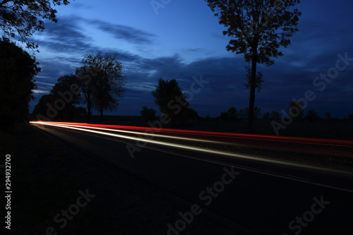 Światła samochodów w nocy na drodze między polami o zmierzchu.