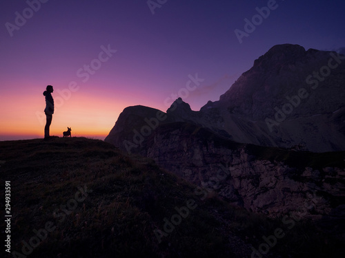 SILHOUETTE: Woman and her dog standing on grassy hill face a summit at sunset.