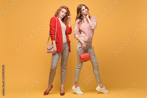 Fashion. Two Beautiful woman in autumn red pink clothes, trendy hair, make up. Adorable well dressed girl, friends on orange. Creative fashionable lady hipster style, beauty colorful shot