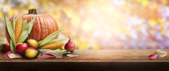 A thanksgiving celebration table of pumpkins, apples, pears and corncobs with autumn sunset foliage in the background.