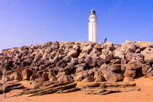 BEACH LANDSCAPE WITH ROCKS ON THE BEACH AND LIGHTHOUSE OF TRAFALGAR ON BLUE SKY. CADIZ BEACHES IN SPAIN