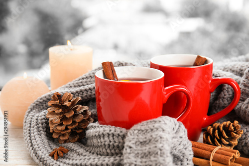Cups of hot winter drink with knitted sweater on window sill indoors