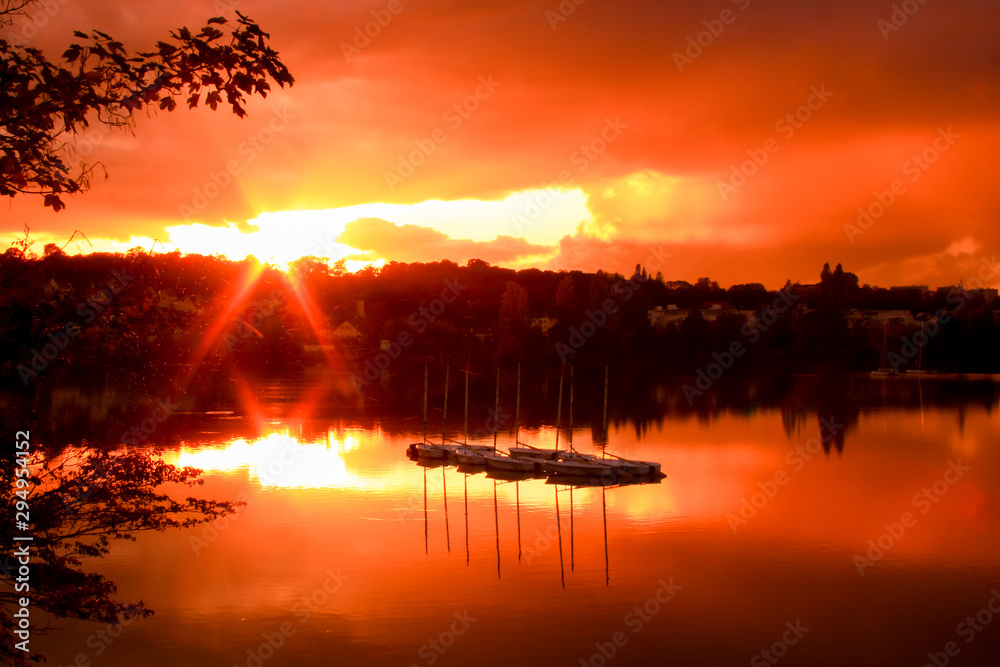 Group of sailboats on a lake at sunset. Houses and forest in the background. Colored dramatic sky and clouds. 