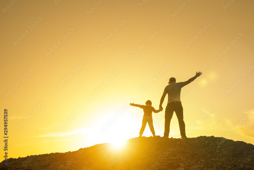 Fototapeta premium Father with a baby girl on top of the mountain, raising his hands up, playing outdoors on a sunset background. Happy loving family