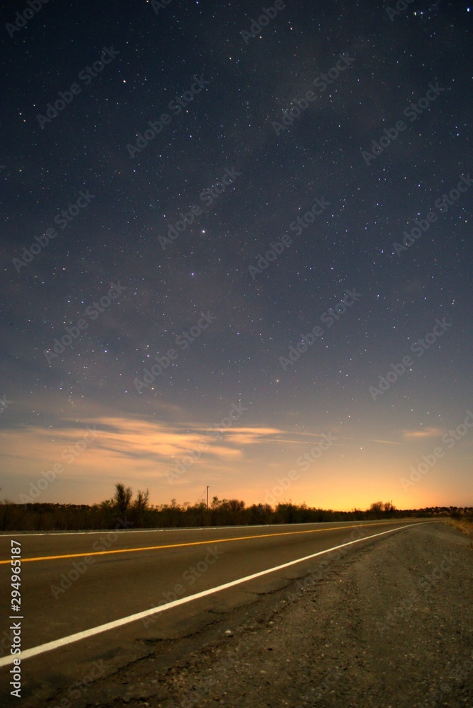 Asphalt road vanishes on the horizon at night under a deep, blue starry sky.