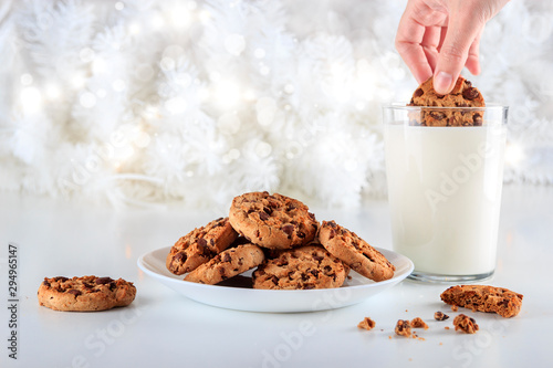 Chocolate chip cookies handmade on a plate. A woman's hand dips a cookie in a glass of fresh milk on Christmas day. The lights are on decorating the white Christmas tree. Christmas holidays concept