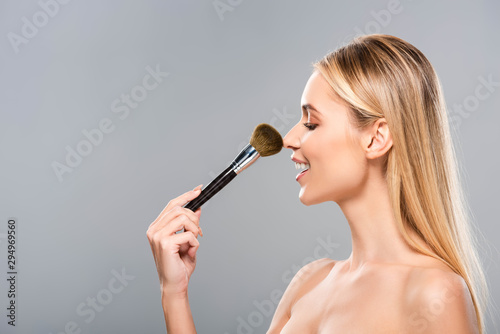 side view of smiling young naked woman with cosmetic brush isolated on grey