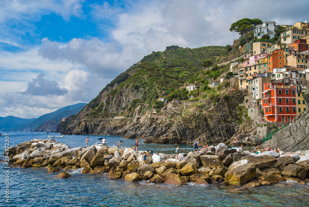 Cinque Terre, Italy - August 17, 2019: Beautiful colorful houses on the rocky coast of the Ligurian Sea. Spreading tree on the mountain. Tourists sunbathe on the rocks