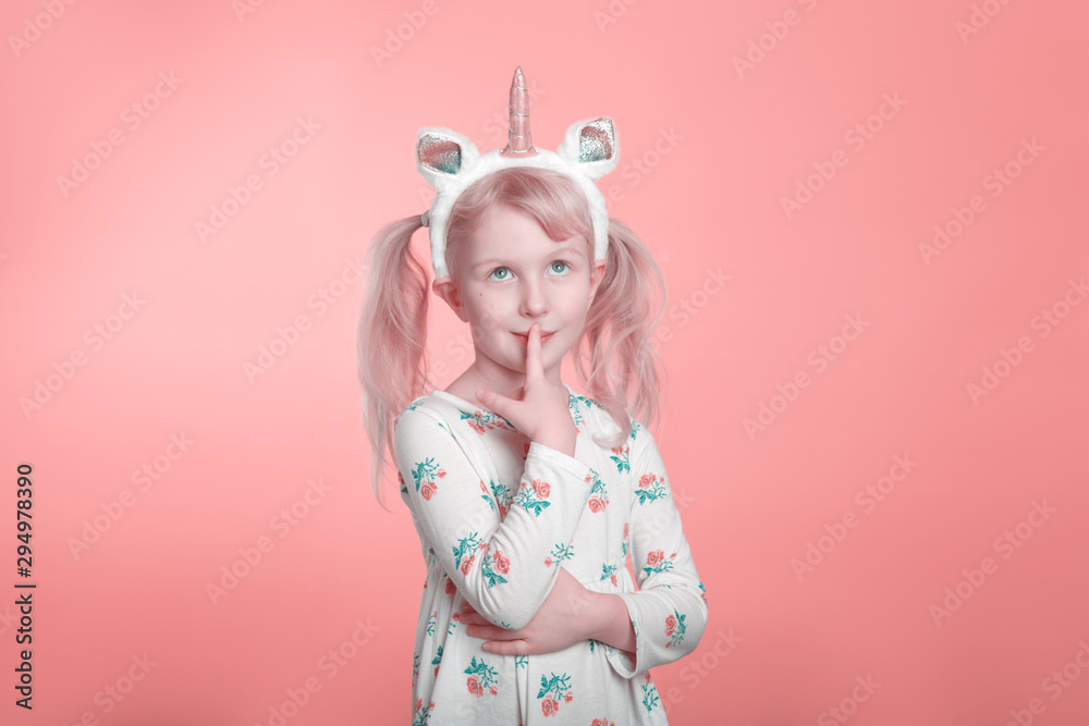 Cute adorable Caucasian blonde girl in white dress wearing unicorn headband horn and ears thinking dreaming. Funny kid child expressing emotion standing in studio on pink coral background.