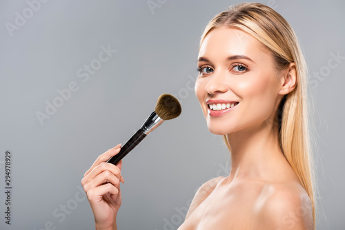 side view of smiling naked woman with cosmetic brush isolated on grey