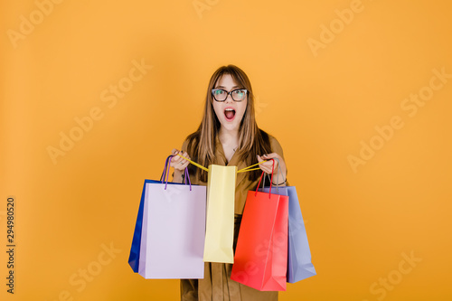 hipster young european woman in glasses and coat with colorful shopping bags isolated over yellow