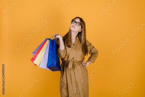 hipster young european woman in glasses and coat with colorful shopping bags isolated over yellow