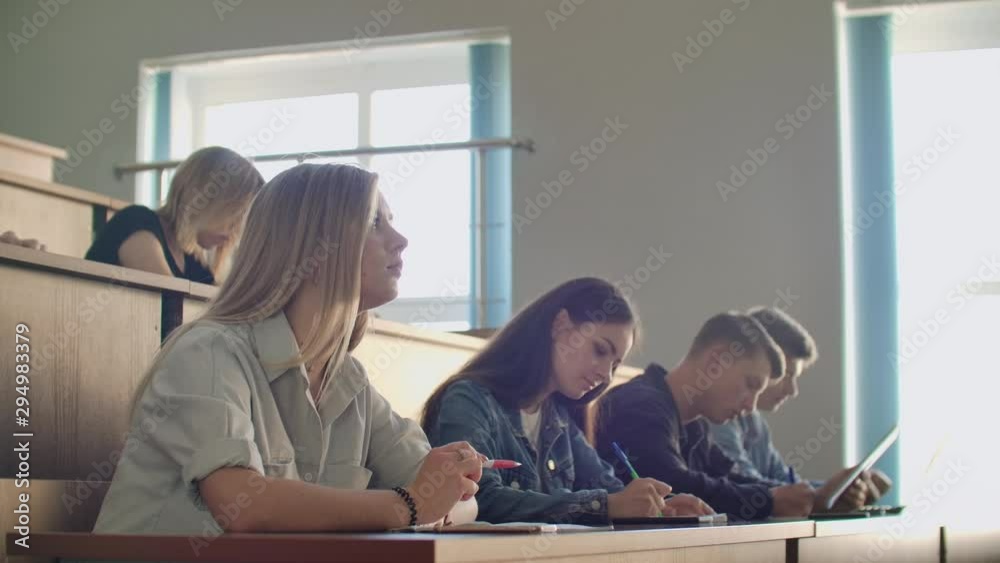 A group of male and female students in a large auditorium of the University listen to a lecture by a Professor