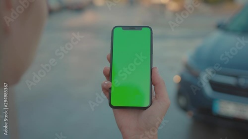 Young woman in close-up holding smartphone watching videos on vertical greenscreen mock-up near parked car outside. Millennial lifestyle.