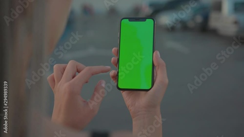 Side view of girl showing modern vertical smartphone in close-up swiping on chrome key greenscreen on blurred background. People in city. Technology.