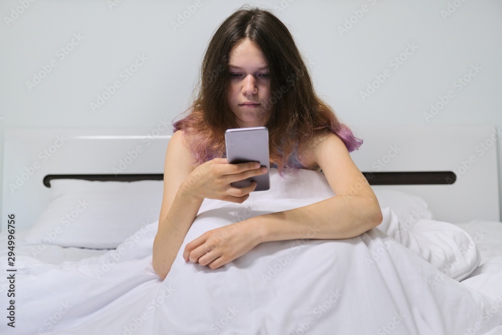 Tired teenager girl sitting at home in bed looking at mobile phone
