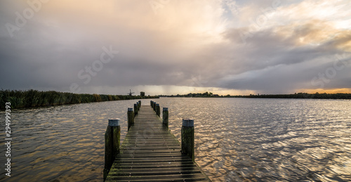 Wooden pier at a lake with windmill and clouds in the distance during sunset