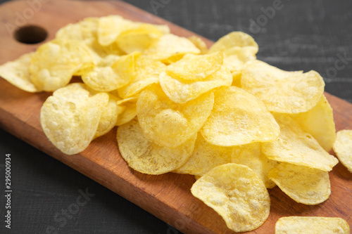 Delicious potato chips with salt on a rustic wooden board on a black surface, side view. Closeup.